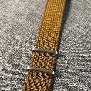 K&ouml;nnte beinhalten: Ein braunes Nylon-Uhrenarmband mit silberner Schlie&szlig;e und Schlaufen.