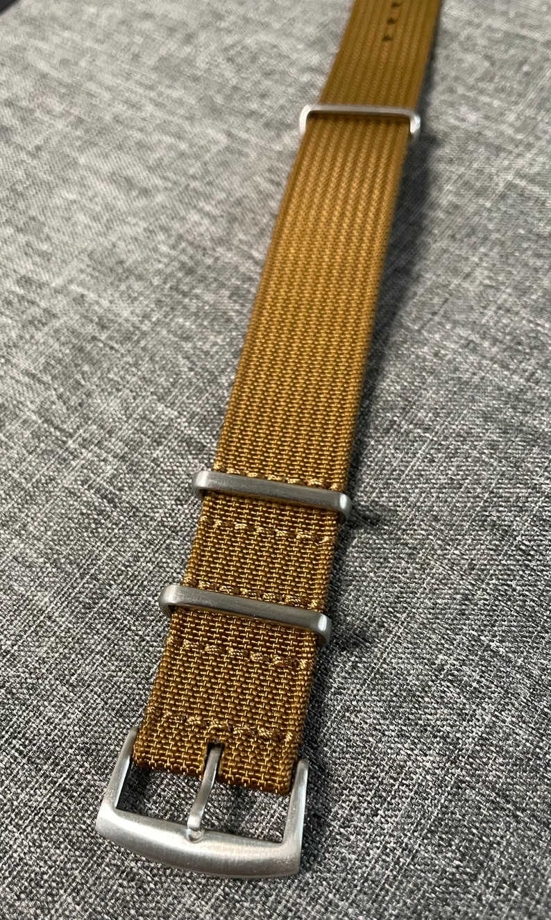 K&ouml;nnte beinhalten: Ein braunes Nylon-Uhrenarmband mit silberner Schlie&szlig;e und Schlaufen.