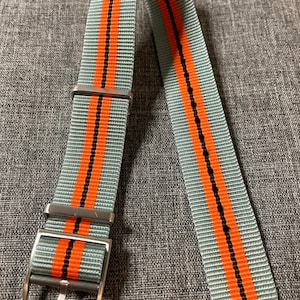 Puede incluir: Una correa de reloj gris con dos rayas naranjas y costuras negras. La correa tiene una hebilla plateada.
