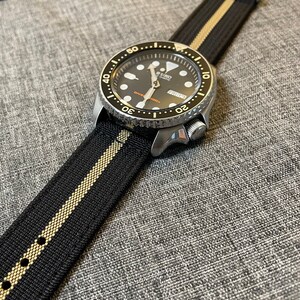 May include: A black and gold striped watch strap with a silver watch face. The watch face has a black background with white markings and a silver second hand. The watch face also has the text "SEIKO AUTOMATIC" and "DIVER'S 200m" printed on it.