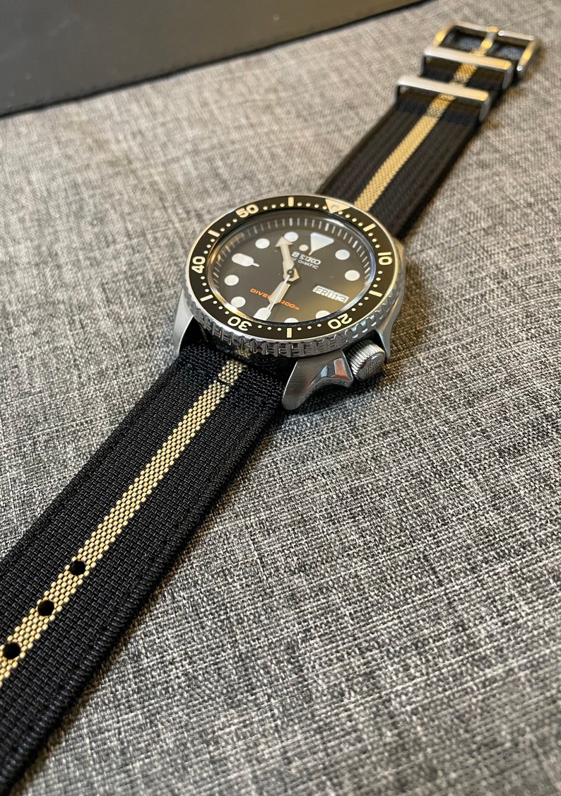 May include: A black and gold striped watch strap with a silver watch face. The watch face has a black background with white markings and a silver second hand. The watch face also has the text "SEIKO AUTOMATIC" and "DIVER'S 200m" printed on it.