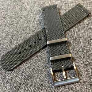 K&ouml;nnte beinhalten: Ein schwarzes Nylon-Uhrenarmband mit einer silbernen Schlie&szlig;e. Das Armband hat eine strukturierte Webart und ist f&uuml;r eine Uhr mit einer Bandansto&szlig;breite von 20 mm ausgelegt.