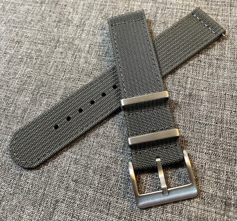 K&ouml;nnte beinhalten: Ein schwarzes Nylon-Uhrenarmband mit einer silbernen Schlie&szlig;e. Das Armband hat eine strukturierte Webart und ist f&uuml;r eine Uhr mit einer Bandansto&szlig;breite von 20 mm ausgelegt.