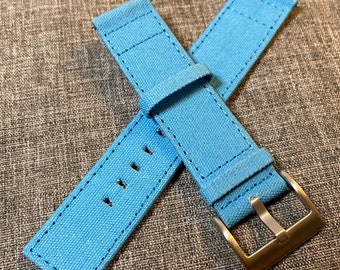 May include: A light blue canvas watch strap with a silver buckle. The strap is made of a soft, durable material and is designed to be comfortable to wear.