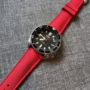 Puede incluir: Un reloj de pulsera con esfera negra, bisel plateado y correa roja brillante. La esfera del reloj tiene agujas e indicadores luminosos, con la marca "Seiko" visible. La correa roja es texturizada y se asegura con una hebilla plateada.