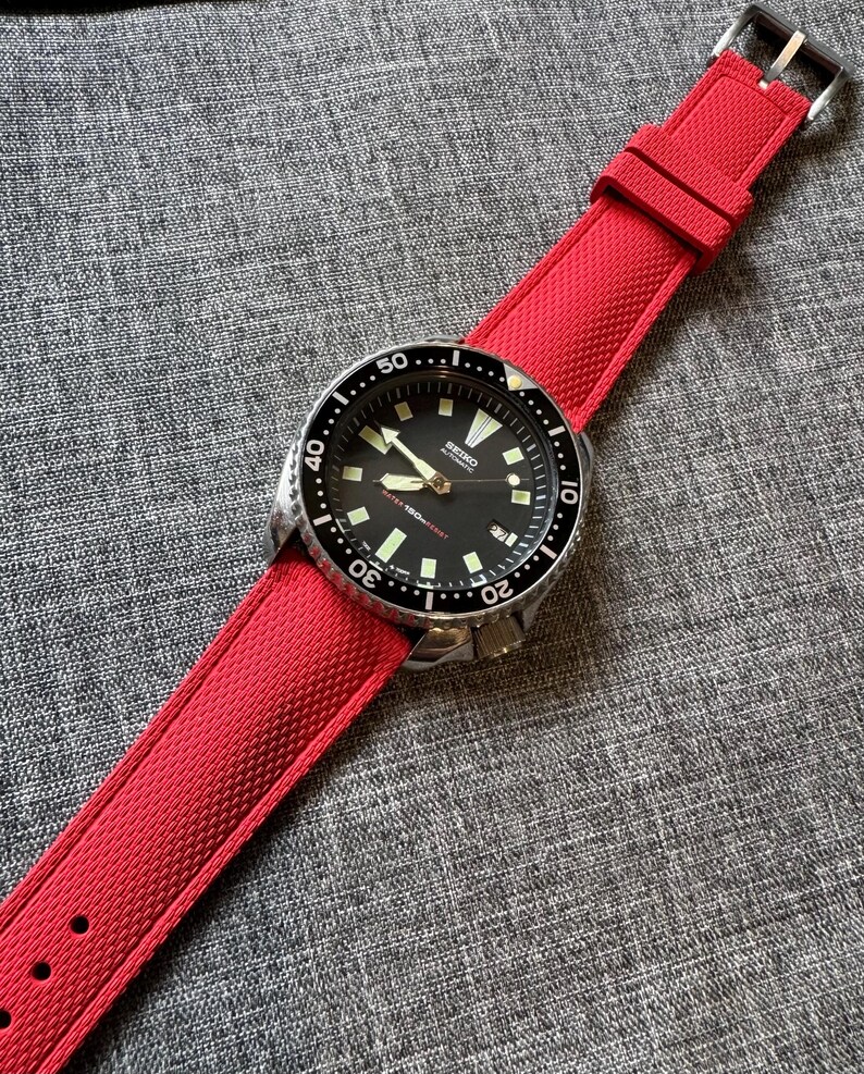 Puede incluir: Un reloj de pulsera con esfera negra, bisel plateado y correa roja brillante. La esfera del reloj tiene agujas e indicadores luminosos, con la marca "Seiko" visible. La correa roja es texturizada y se asegura con una hebilla plateada.