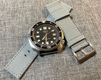May include: A black-faced Seiko automatic watch with a silver bezel and a gray canvas strap. The watch face has white markers and hands, with the date displayed at the 3 o'clock position. The strap has a silver buckle.
