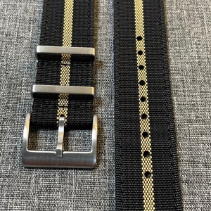 May include: A black and gold striped watch strap with a silver buckle. The strap is made of nylon and has a textured finish.