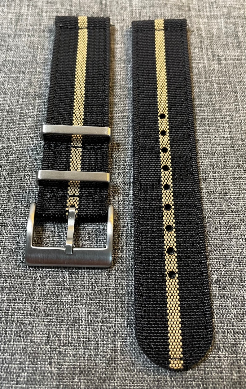 May include: A black and gold striped watch strap with a silver buckle. The strap is made of nylon and has a textured finish.