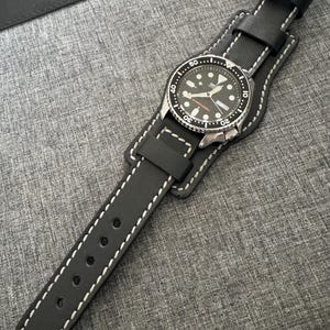 May include: A black wristwatch with a black leather strap and white stitching. The watch face is black with white and silver details. The watch is attached to a wider black leather strap.