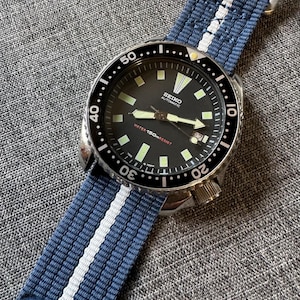 Peut inclure: Une montre-bracelet automatique Seiko avec un cadran noir et un bracelet en nylon à rayures bleues et blanches. La montre a un boîtier en acier inoxydable et une lunette rotative. Le cadran affiche l'heure et la date. La montre est étanche à 150m.