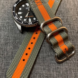 May include: A gray and orange striped nylon watch strap with silver hardware. The strap has two keepers and a buckle.