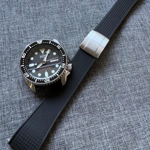 May include: A black and silver diver's watch with a black rubber strap. The watch face has white markings and a date display. The strap has a textured pattern and a silver buckle. The watch is on a gray surface.