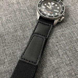 May include: A black and silver wristwatch with a black nylon strap and a Union Jack design on the strap. The watch face is black with white markings and a date window.
