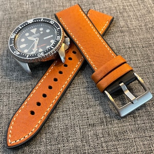 May include: A brown leather watch strap with white stitching and a silver buckle. The strap is shown with a black watch face with silver hands and markers.