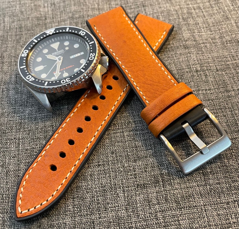 May include: A brown leather watch strap with white stitching and a silver buckle. The strap is shown with a black watch face with silver hands and markers.