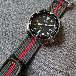 May include: A black-faced Seiko automatic watch with a gray and red striped nylon strap. The watch has a stainless steel case and a rotating bezel. The strap is approximately 22 mm wide.