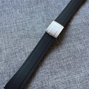 Puede incluir: Una correa de reloj negra con una hebilla plateada. La correa est&aacute; hecha de un material flexible y tiene una hebilla rectangular con una superficie texturizada. La correa est&aacute; sobre una superficie gris texturizada.