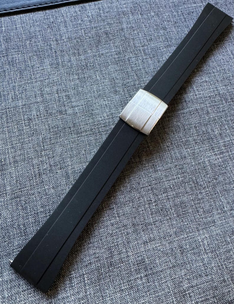 Puede incluir: Una correa de reloj negra con una hebilla plateada. La correa est&aacute; hecha de un material flexible y tiene una hebilla rectangular con una superficie texturizada. La correa est&aacute; sobre una superficie gris texturizada.