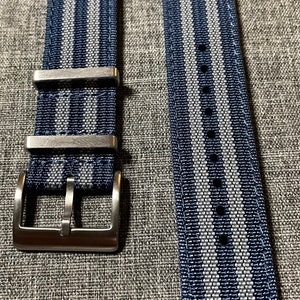 May include: A blue and gray striped watch strap with a silver buckle. The strap is made of woven nylon and has a textured finish.