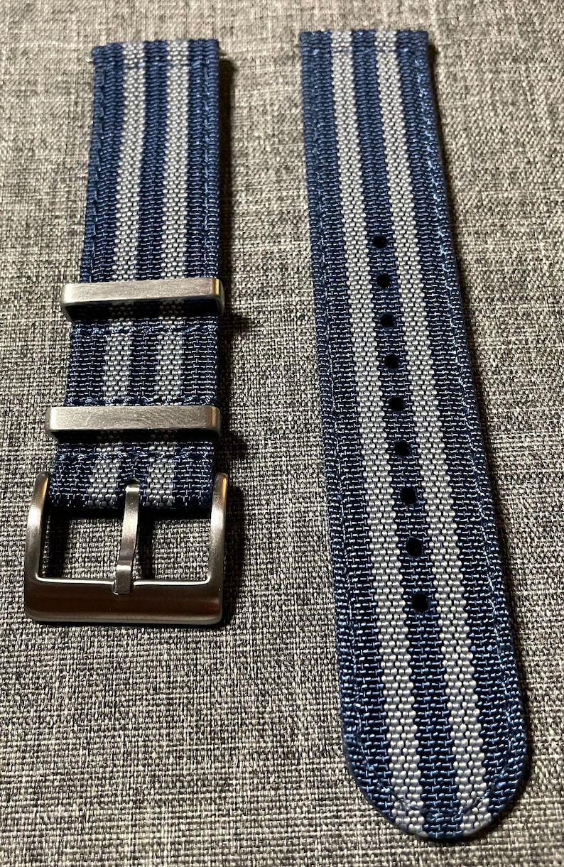 May include: A blue and gray striped watch strap with a silver buckle. The strap is made of woven nylon and has a textured finish.