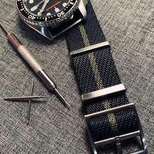 May include: A black and silver watch with a black nylon watch strap with green stripes. The watch face has white markings and the text "Diver's 200m".
