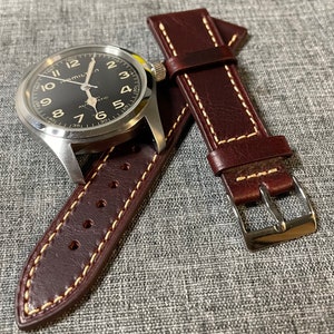 Può includere: Un cinturino da orologio in pelle marrone con cuciture bianche. Il cinturino ha una fibbia ed è mostrato con un orologio argentato con un quadrante nero.
