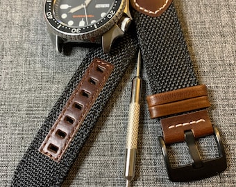 May include: A black and silver wristwatch with a black dial and a rotating bezel, alongside a black nylon watch strap with brown leather accents and a metal tool. The watch strap has a black buckle and brown leather details.