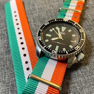 May include: A black Seiko Automatic watch with a green, white and orange striped nylon watch strap. The watch face has a black background with white markings and a date window at the 3 o'clock position. The watch is water resistant to 150 metres.