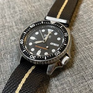 May include: A black Seiko automatic diver's watch with a black and white striped nylon strap. The watch face has a black background with white markings and a date window that reads "TUE 16". The bezel is black with white markings and reads "50... 20".