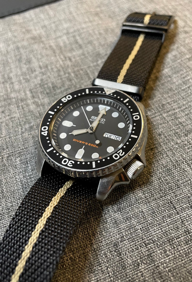 May include: A black Seiko automatic diver's watch with a black and white striped nylon strap. The watch face has a black background with white markings and a date window that reads "TUE 16". The bezel is black with white markings and reads "50... 20".