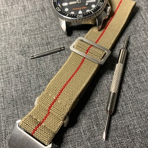 May include: A close-up of a beige watch strap with a red stripe, a silver buckle, and two spring bars. The strap is lying on a gray textured surface.