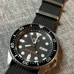 May include: A black Seiko automatic diver's watch with a black nylon strap. The watch face has white markings and a date window that reads "MON 20". The bezel is black with white markings and reads "Diver's 200m".