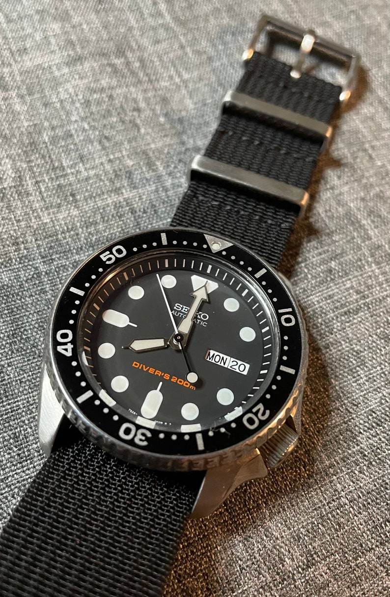 May include: A black Seiko automatic diver's watch with a black nylon strap. The watch face has white markings and a date window that reads "MON 20". The bezel is black with white markings and reads "Diver's 200m".