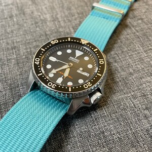 May include: A black and silver Seiko automatic watch with a blue nylon strap. The watch face has a black background with white markings and a date window that reads "FRI 13".