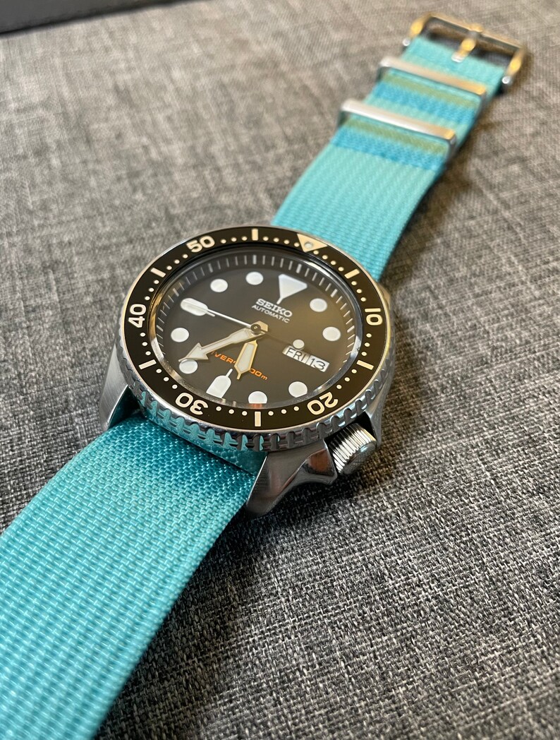 May include: A black and silver Seiko automatic watch with a blue nylon strap. The watch face has a black background with white markings and a date window that reads "FRI 13".