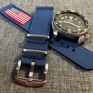 Op de afbeelding: Een blauwe nylon horlogeband met een stalen gesp met een klein Amerikaans vlaggetje. De band wordt getoond met een zilveren horloge met een zwarte wijzerplaat en een draaibare bezel.