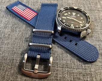 May include: A blue nylon watch strap with a stainless steel buckle featuring a small American flag. The strap is shown with a silver watch with a black face and a rotating bezel.