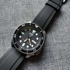 May include: A black Seiko automatic wristwatch with a black bezel and a black rubber strap. The watch face is black with white hour markers and hands. The watch has a date display and is water resistant to 200m.