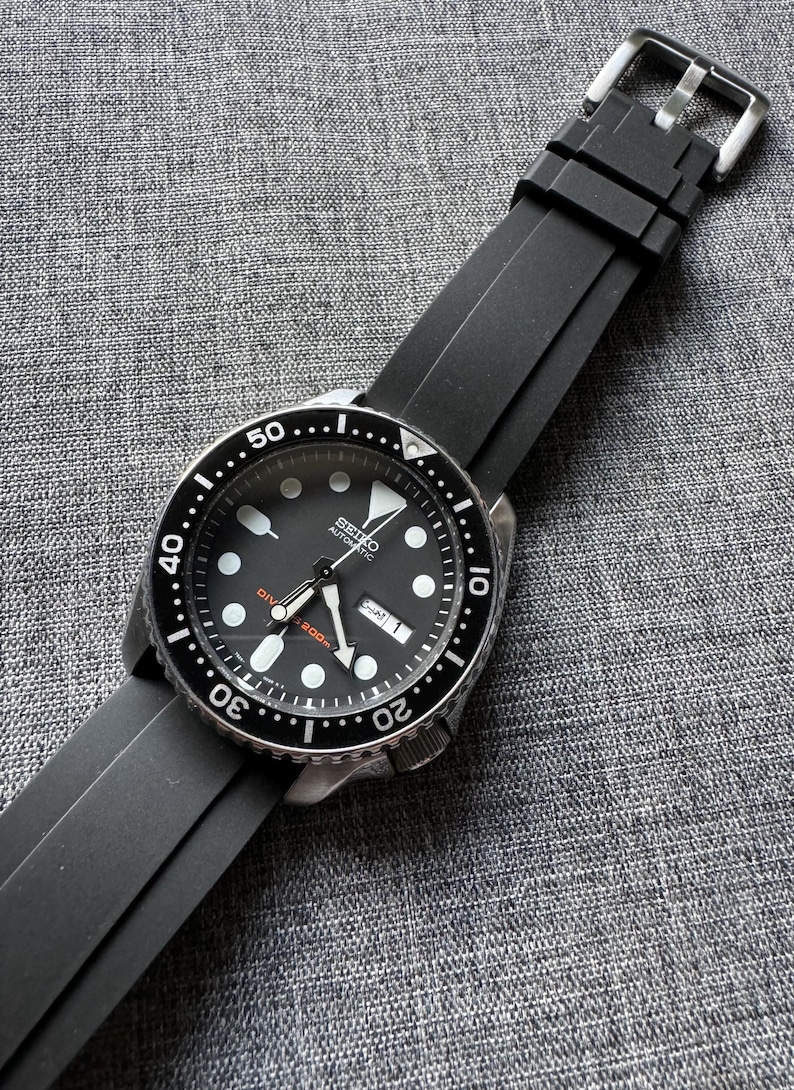 May include: A black Seiko automatic wristwatch with a black bezel and a black rubber strap. The watch face is black with white hour markers and hands. The watch has a date display and is water resistant to 200m.