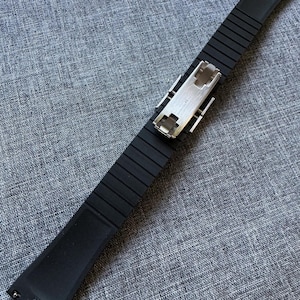 Puede incluir: Una correa de reloj negra con un dise&ntilde;o acanalado y un cierre plateado. La correa est&aacute; hecha de un material flexible y est&aacute; en &aacute;ngulo sobre una superficie gris texturizada. La correa mide aproximadamente 2,7 cm de ancho.