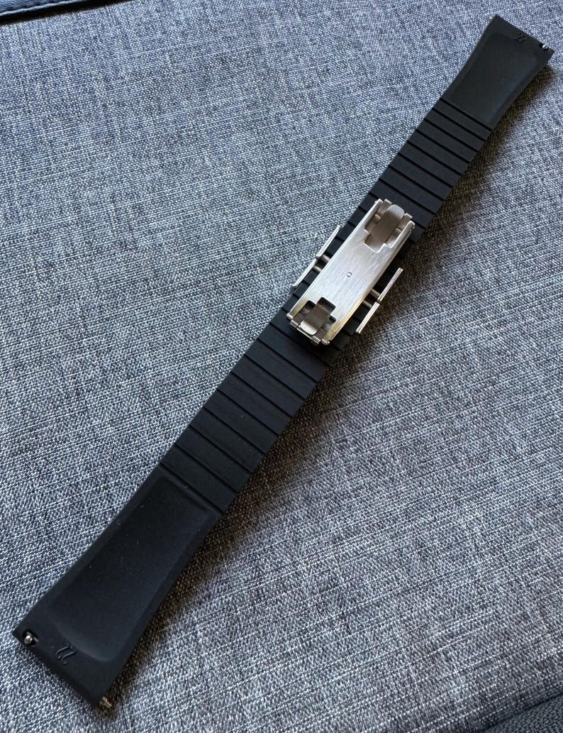 Puede incluir: Una correa de reloj negra con un dise&ntilde;o acanalado y un cierre plateado. La correa est&aacute; hecha de un material flexible y est&aacute; en &aacute;ngulo sobre una superficie gris texturizada. La correa mide aproximadamente 2,7 cm de ancho.