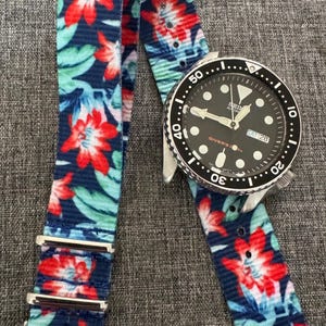May include: A wristwatch with a black face and a silver bezel, paired with a watch strap adorned with a floral pattern. The strap showcases red flowers and green leaves against a navy blue backdrop. The watch shows the day and date.
