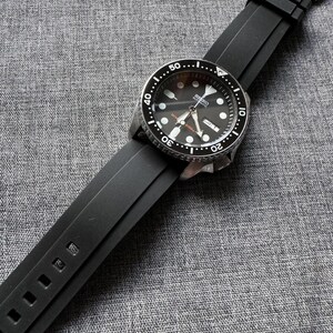 May include: A black Seiko automatic wristwatch with a black dial and a black rubber strap. The watch features a silver bezel with white markings and a date display. The strap is secured with a silver buckle.