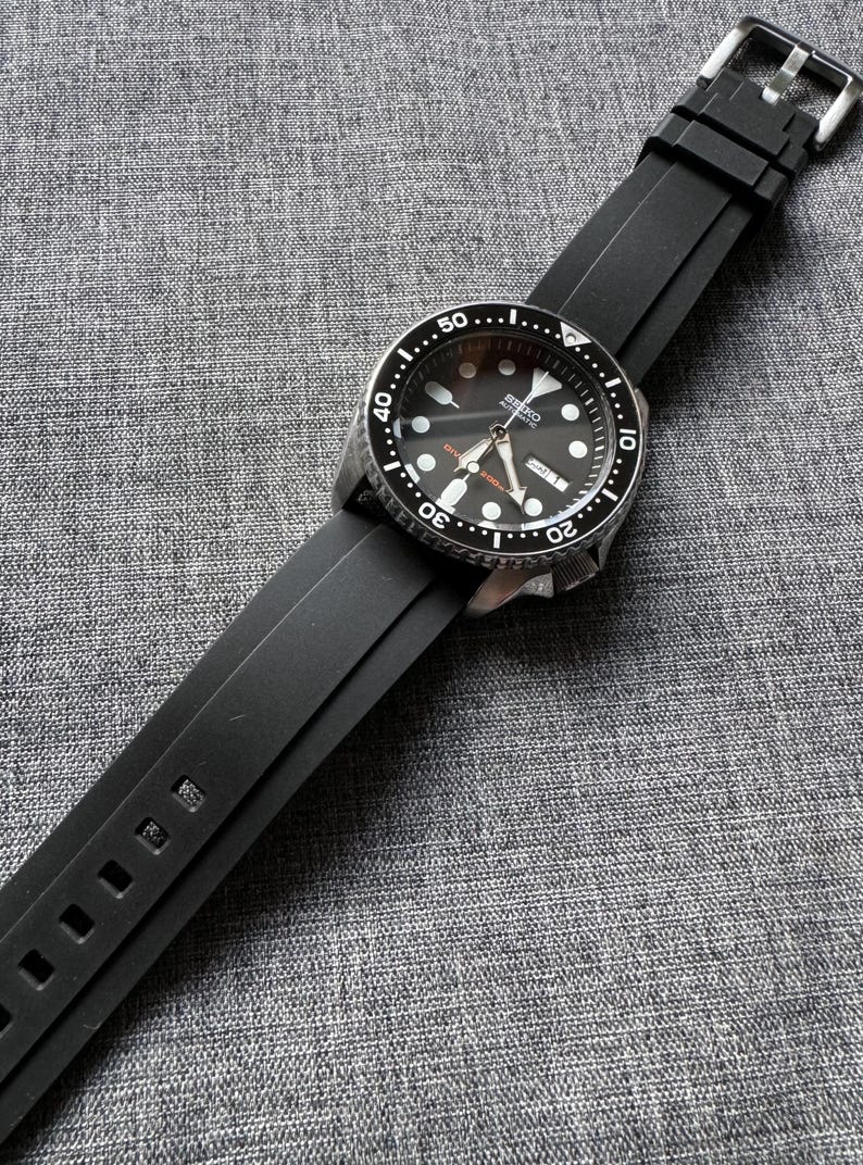 May include: A black Seiko automatic wristwatch with a black dial and a black rubber strap. The watch features a silver bezel with white markings and a date display. The strap is secured with a silver buckle.