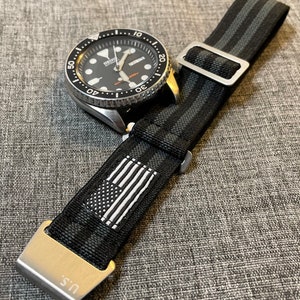 以下が含まれることがあります： 黒とグレーのストライプの腕時計ストラップで、銀色のバックルと小さなアメリカ国旗のパッチが付いています。ストラップはナイロン製で、質感のある仕上げになっています。ストラップは、ラグ幅が20mmの腕時計に合うように設計されています。