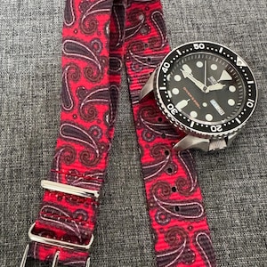 May include: A wristwatch with a black face and a silver case, paired with a red paisley patterned watch strap. The strap has a repeating pattern of dark red and brown paisley designs on a bright red background. The watch face shows the day and date.