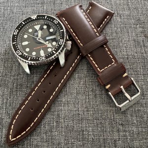 May include: A black-faced wristwatch with a silver bezel and a brown leather watch strap. The watch face displays the day and date. The watch strap is brown with white stitching and a silver buckle. The watch is a diver's watch.