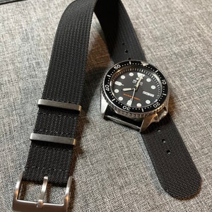 May include: A black watch with a black nylon strap. The watch face is black with white markings and a date window. The watch has a silver bezel with white markings. The watch is on a gray surface.