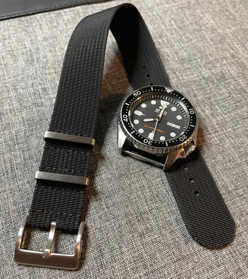 May include: A black watch with a black nylon strap. The watch face is black with white markings and a date window. The watch has a silver bezel with white markings. The watch is on a gray surface.